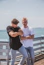 Two men laughing and posing outdoors against a sea backdrop, exuding stylish vibes.